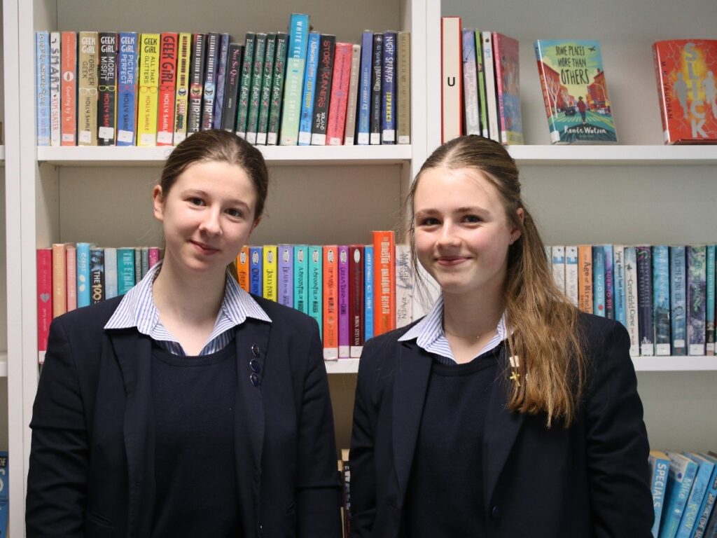 2 student girls stood in front of a bookcase
