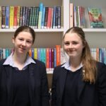 2 student girls stood in front of a bookcase