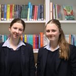 2 student girls stood in front of a bookcase